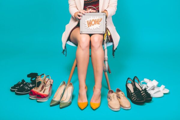 A woman is sitting in a chair, surrounded by a box and an assortment of shoes. The scene showcases various types of footwear, including sandals and high heels, emphasizing fashion and style. The woman's legs are visible, adding to the overall aesthetic of the image, which celebrates clothing and fashion accessories.