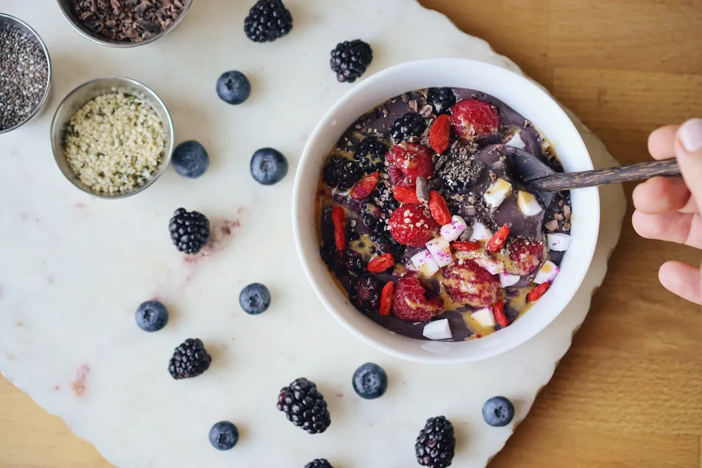 Acai bowl topped with raspberries, blackberries, blueberries, goji berries, chia seeds, and coconut, served on a marble board with scattered fresh berries.