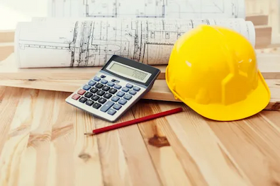 Yellow construction helmet, calculator, red pencil, and blueprints on a wooden table.